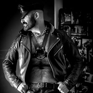 Photo of Joe King in black & white wearing an open leather biker jacket and leather harness